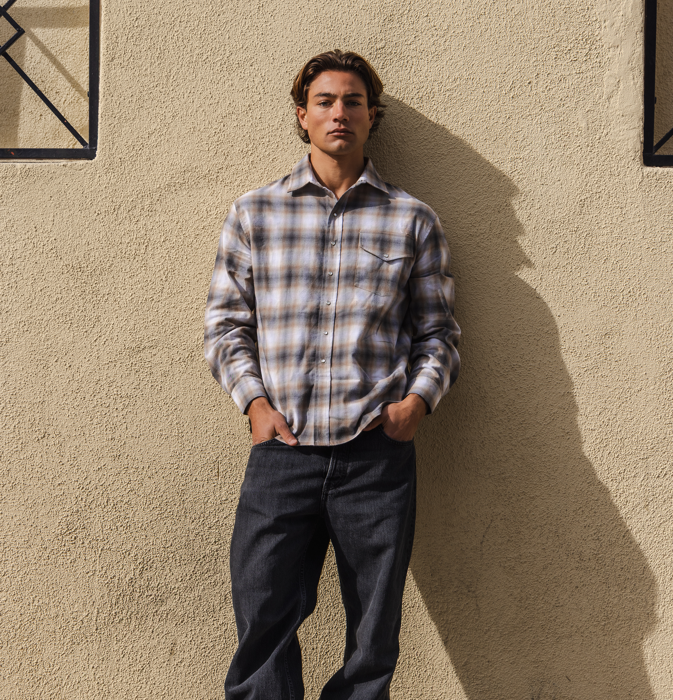 Man wearing a plaid shirt and dark jeans standing against a beige wall.
